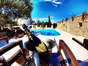 Zona junto a la piscina con mobiliario de mimbre, una botella de champán en un cubo y un muro de piedra.