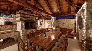 Uma área de refeições rústica com uma mesa de madeira comprida e cadeiras, paredes de pedra e um forno a lenha.