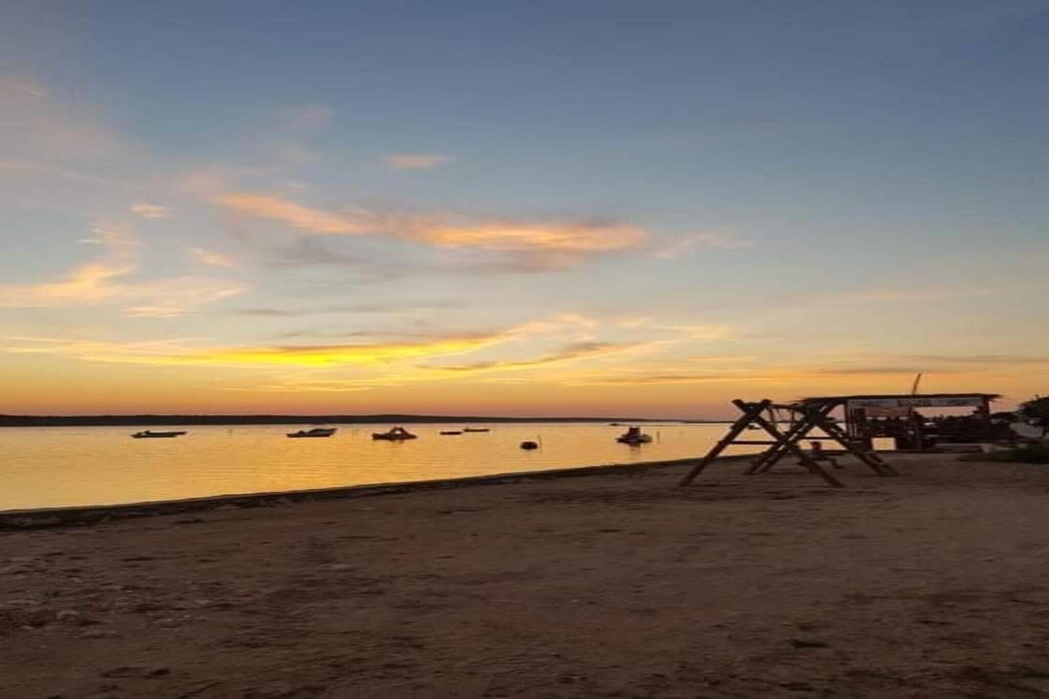 Una playa con una estructura de madera, posiblemente un sillón o una pequeña mesa, y varios barcos en aguas tranquilas durante el atardecer.