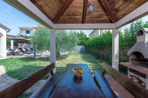 Une pergola en bois, avec une table et un saladier rempli de fruits au centre, donnant sur un jardin avec une piscine et des chaises longues.