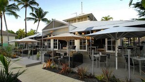 An outdoor dining area with tables and umbrellas, surrounded by palm trees and a building with a sloped roof.