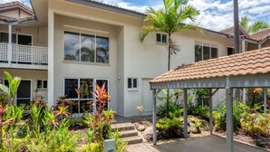 A modern two-story building with a covered patio area, surrounded by tropical plants and a small garden.
