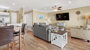 A living room with a grey sofa, a white armchair, a wooden dining table, and a ceiling fan. - Tybeedaze at the Beach! | Heated Pool Access (Tybee Island)