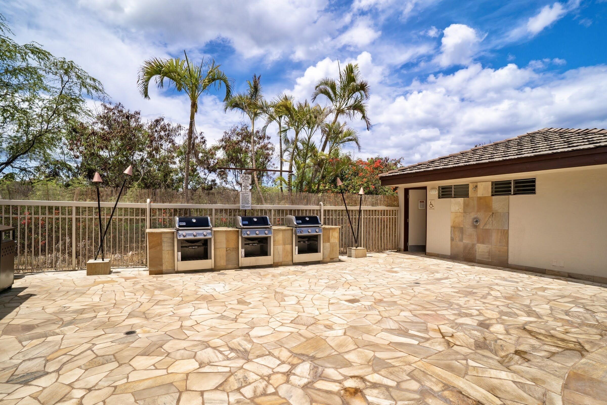 An outdoor patio with stone flooring, a barbecue area with four grills, and a building with a tiled roof.