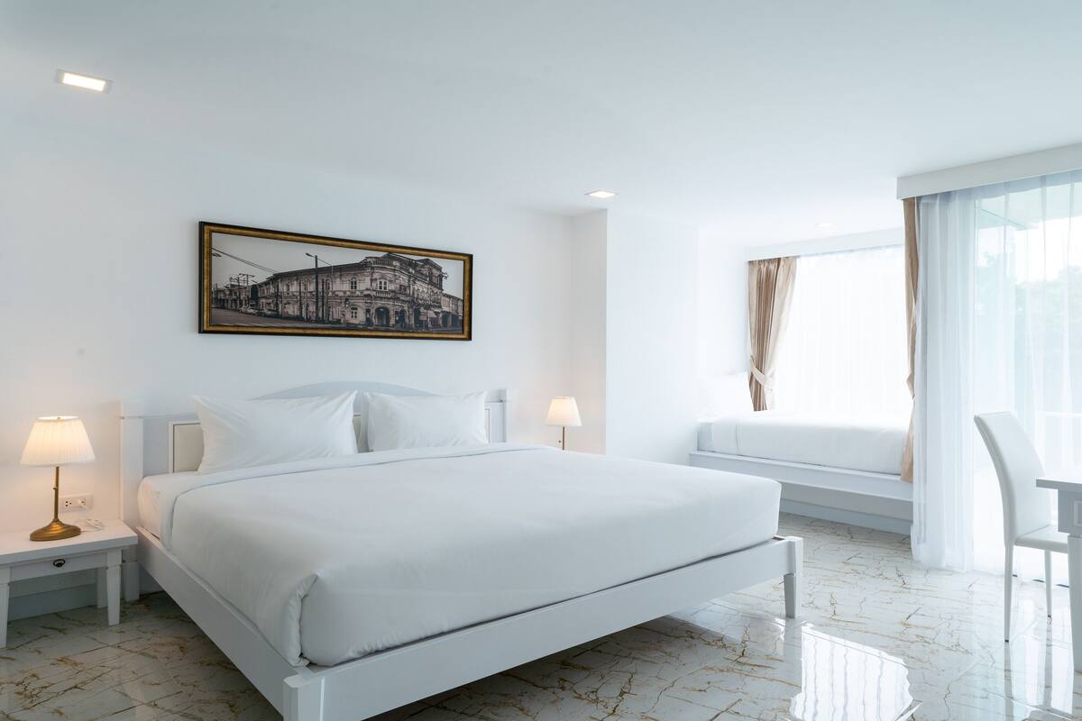 A hotel room with a large bed, bedside lamps, a window with curtains, and a cityscape painting on the wall.
