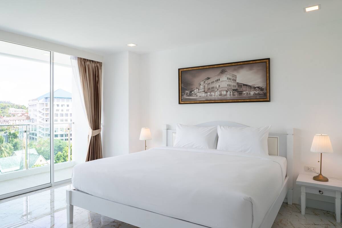 A hotel room with a large bed, two bedside tables with lamps, a painting on the wall, and a view of buildings through large windows.