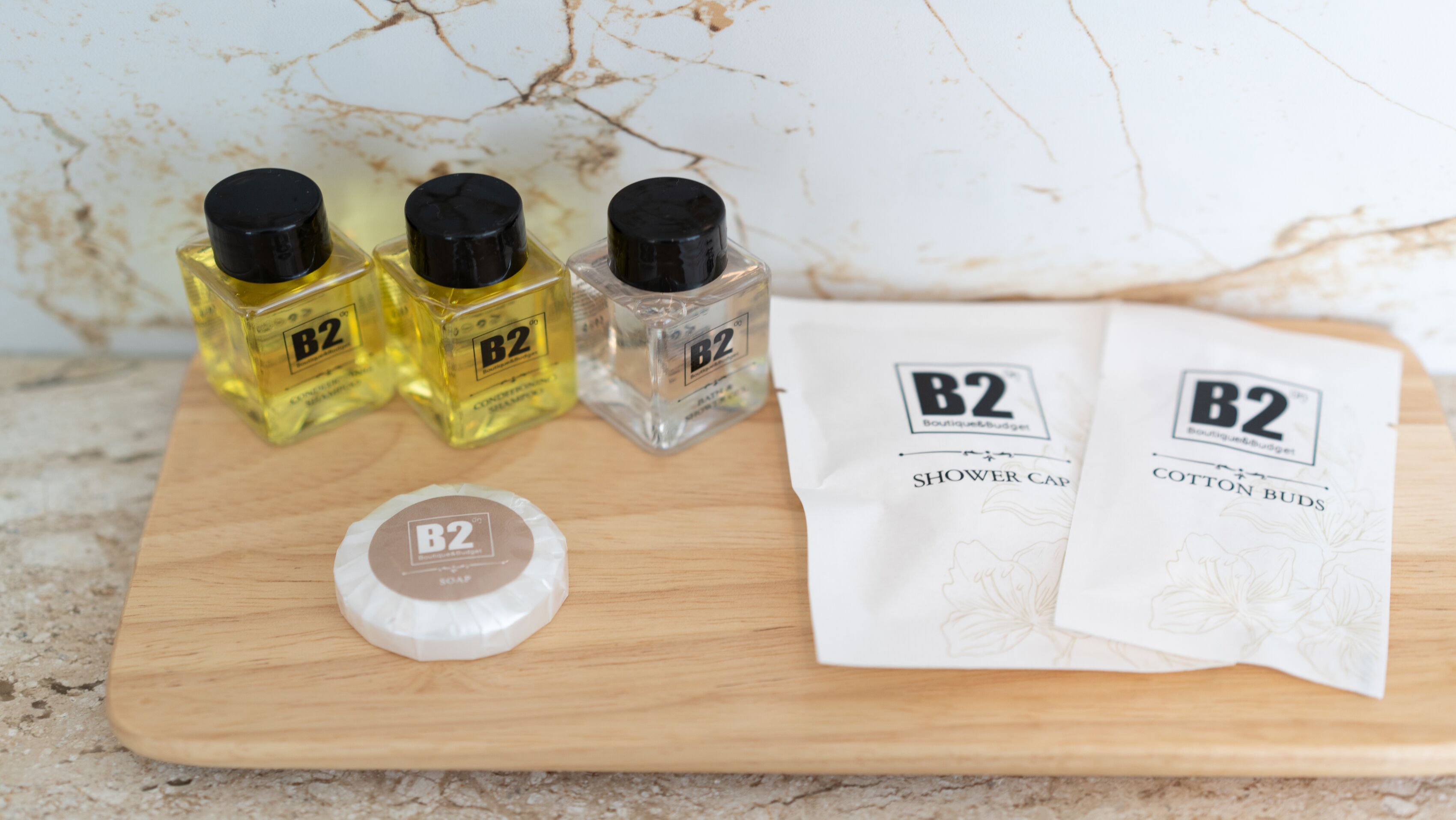 A wooden board with three B2 branded bottles, a shower cap, and two B2 branded cotton bud packets.