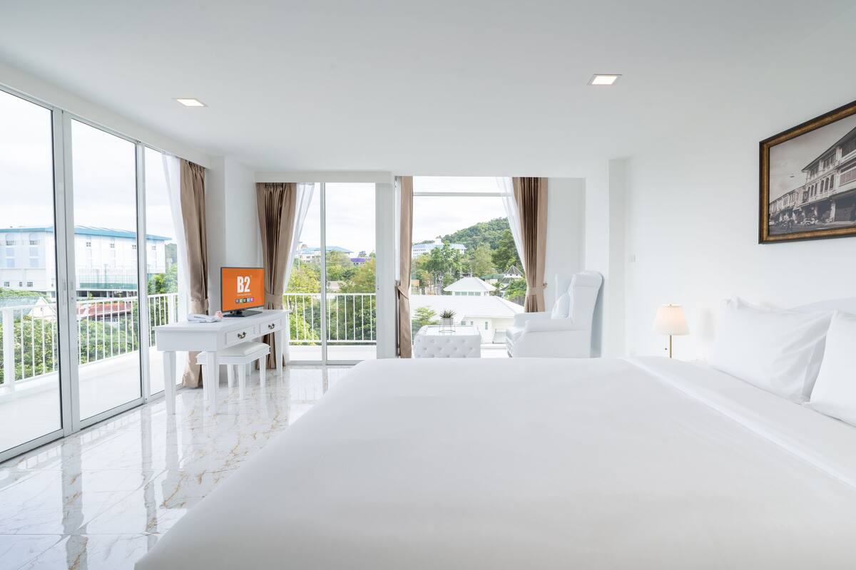 A hotel room with a large bed, a desk with a computer, a chair, and a balcony with a view of buildings and trees.