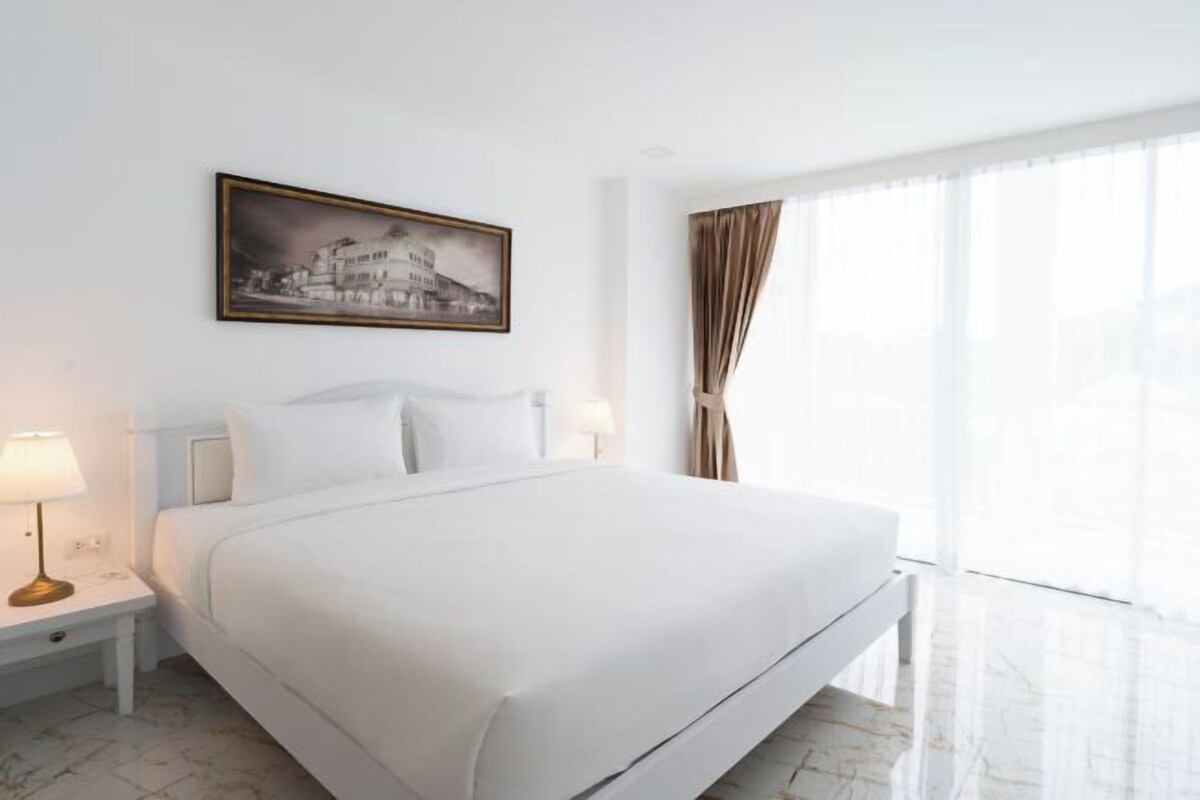 Una habitación de hotel con una cama grande, una mesita de noche con lámpara, un cuadro enmarcado en la pared y un ventanal con cortinas.