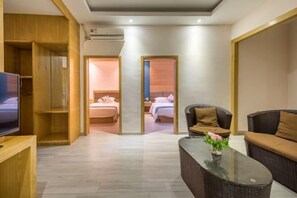 Quarto de hotel moderno com televisão, uma mesa de centro de vidro com um vaso de flores e uma área de estar confortável com sofá e poltrona.