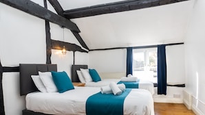 A room with two beds, wooden beams, white walls, and a window with blue curtains. - 2 Pound cottage (Gloucester)