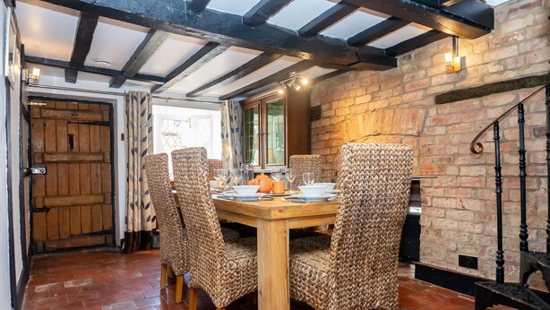 A dining area with a wooden table and wicker chairs, a brick wall, and a wooden door. - 2 Pound cottage (Gloucester)