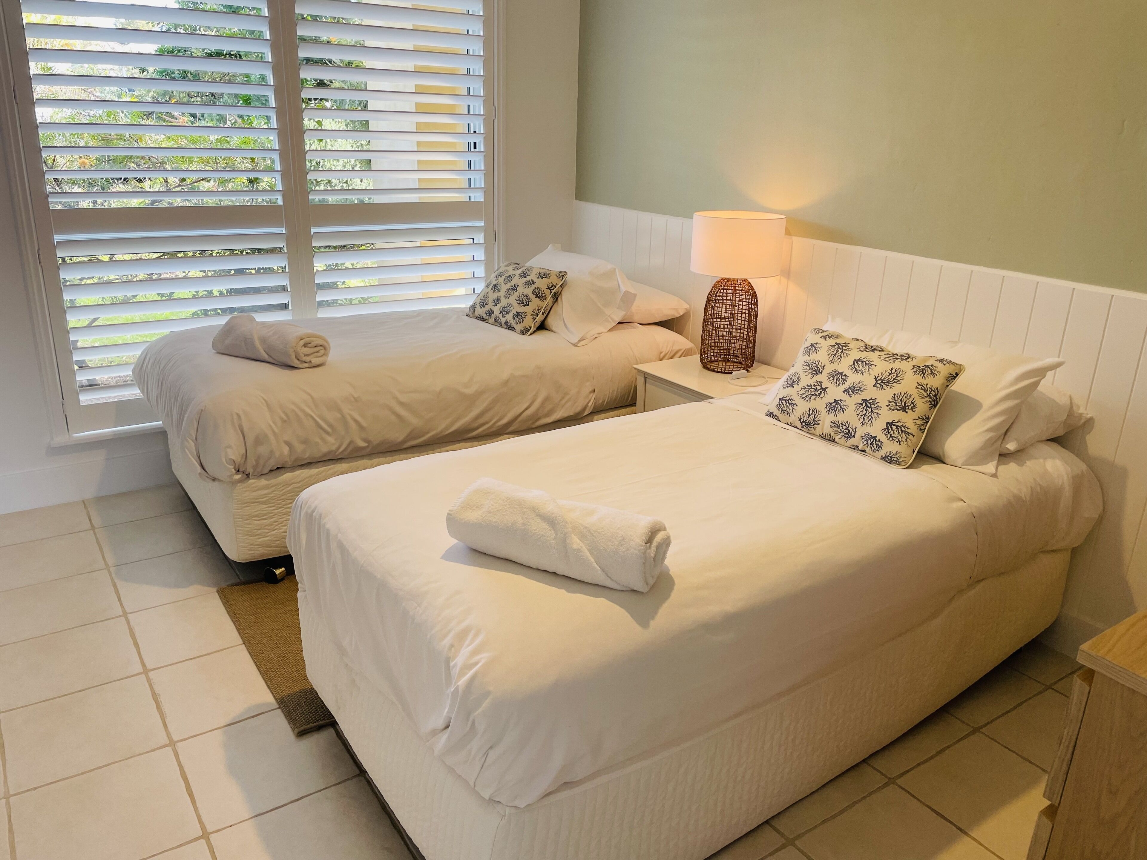 A hotel room with two beds, a window with blinds, a bedside lamp, and a woven nightstand.