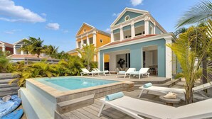 Un'area piscina con lettini e vasca idromassaggio, circondata da piante tropicali e un edificio di due piani con facciata azzurra e gialla.