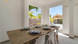 Zona pranzo con tavolo di legno apparecchiato per quattro persone, sedie bianche e vista su una piscina e palme all'esterno.