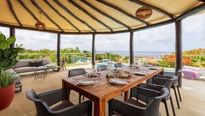 Un patio al aire libre con una mesa de madera dispuesta para comer, rodeada de sillas de mimbre, bajo un toldo a rayas, con vista a una piscina y al océano.