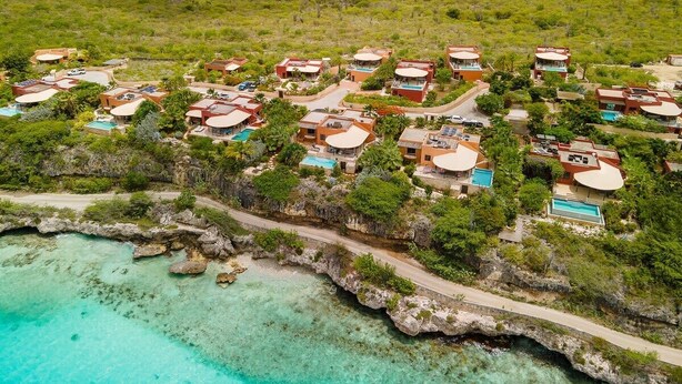 Vista aérea de un complejo turístico costero con varias edificaciones, piscinas y un mar turquesa cristalino.