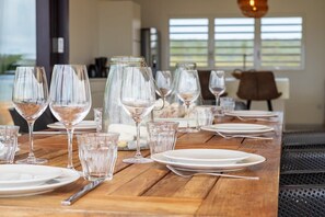 A dining table set with wine glasses, plates, and cutlery, with a view of the outdoors through large windows. - Villa Gemini with Private Pool and Sea View (Kralendijk)