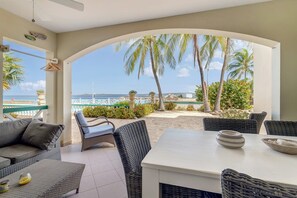 A covered outdoor seating area with wicker furniture, a white table, and a view of the sea and palm trees.