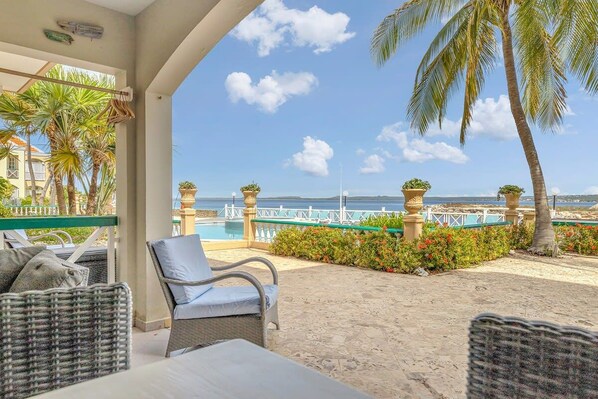 A covered outdoor seating area with wicker furniture, a view of the sea, and palm trees.