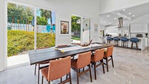 Une cuisine moderne avec des meubles blancs et un îlot central, un coin repas avec une longue table et des chaises orange, et de grandes portes-fenêtres coulissantes donnant sur un jardin.