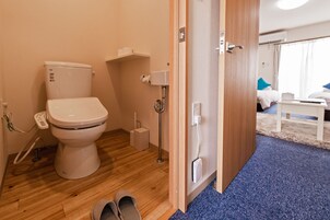 Egy modern fürőszoba, amelyben WC, bidé és zuhany található. Egy faajtó vezet egy hálószobába, ahol egy ágy és egy éjjeliszekrény van.