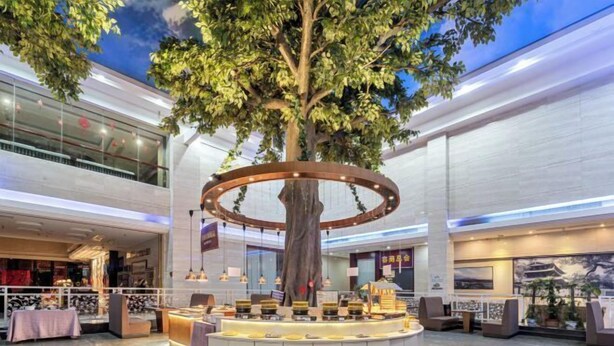A modern indoor atrium with a central tree, seating areas, and retail spaces. - Ronzou International Hotel (Rong County)