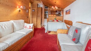 Una habitación acogedora con paredes y techo de madera, una alfombra roja, dos camas de madera con almohadas, una pequeña mesa de comedor con mantel blanco, y un área de cocina visible a través de una puerta abierta.
