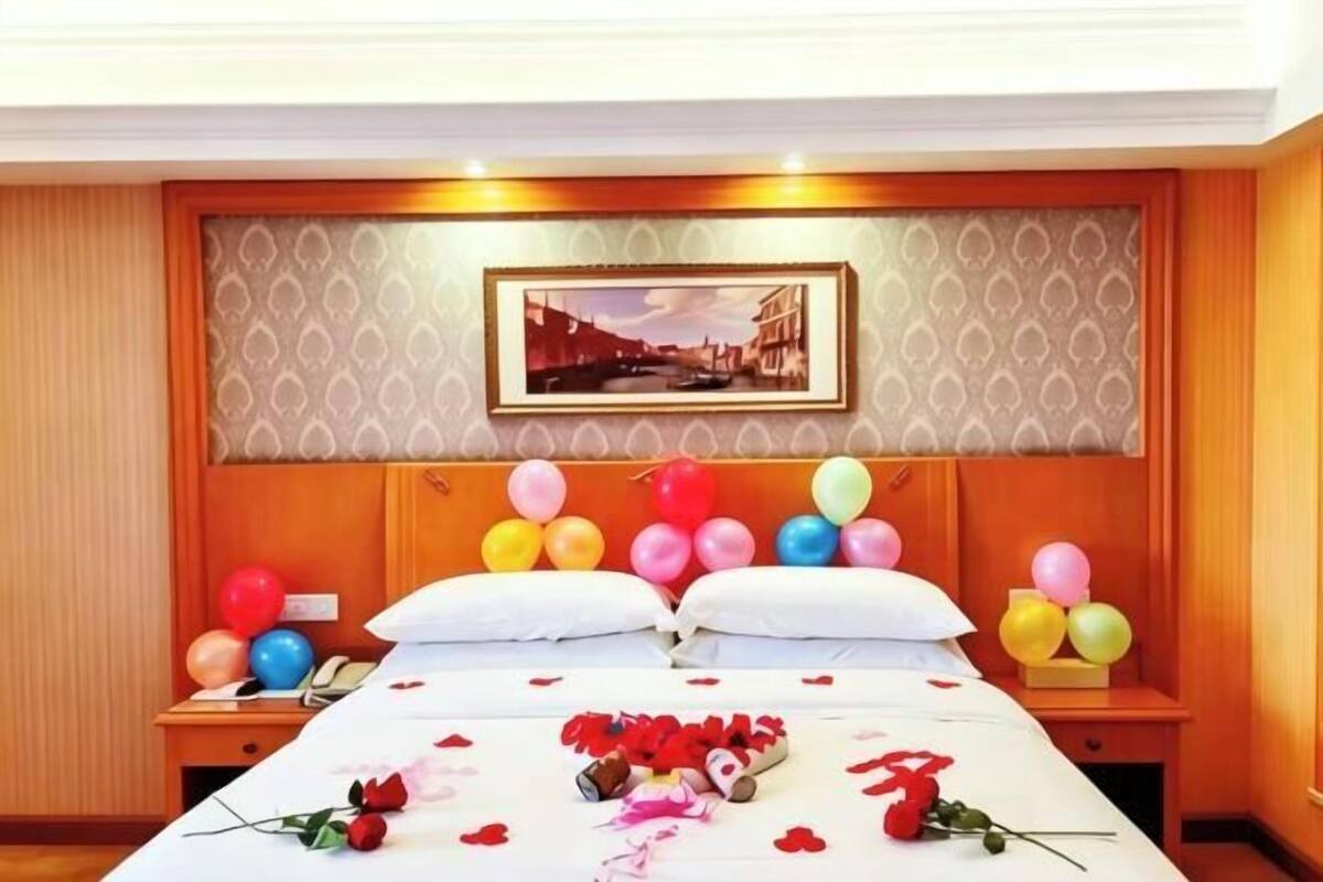 A hotel room with a bed, pillows, a nightstand, a framed picture on the wall, and balloons.