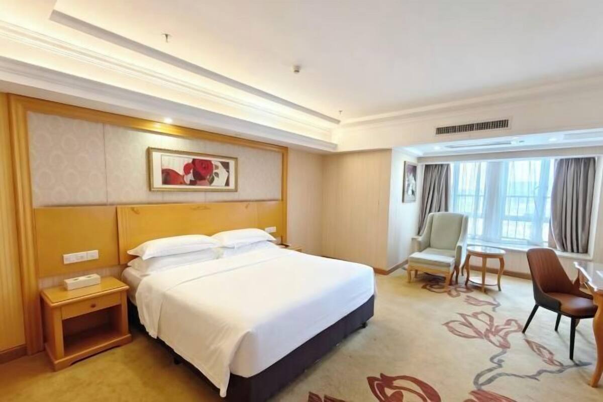 A hotel room with a large bed, a bedside table, a chair, a desk, and a window with curtains.