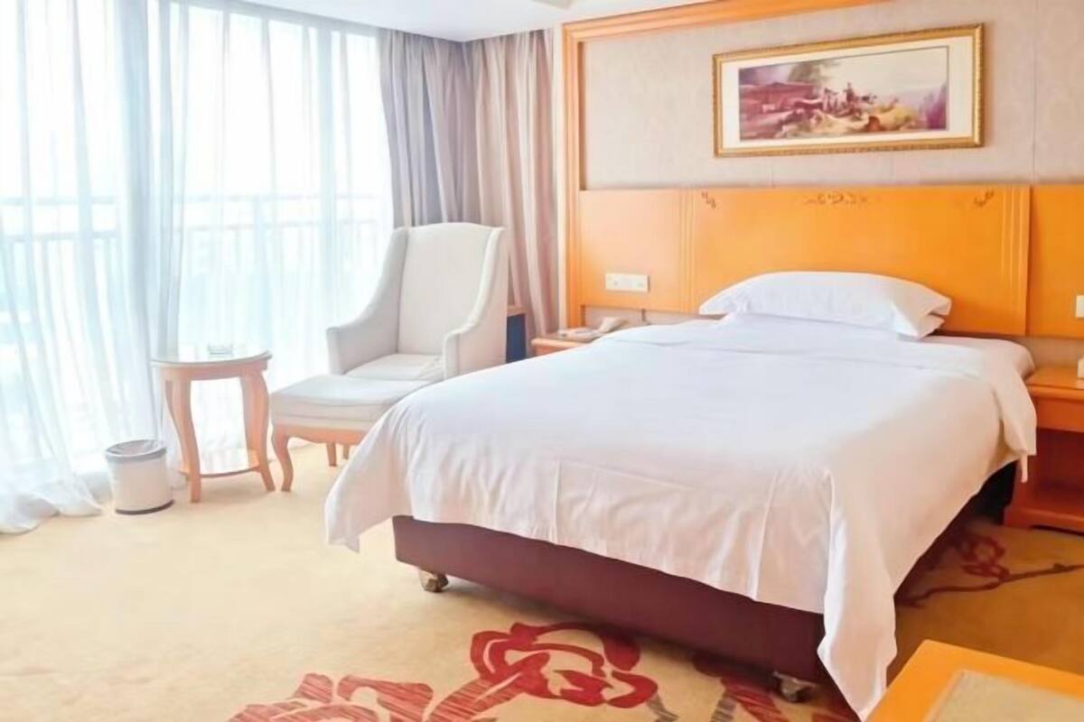 A hotel room with a large bed, a chair, a small table, and a painting on the wall.