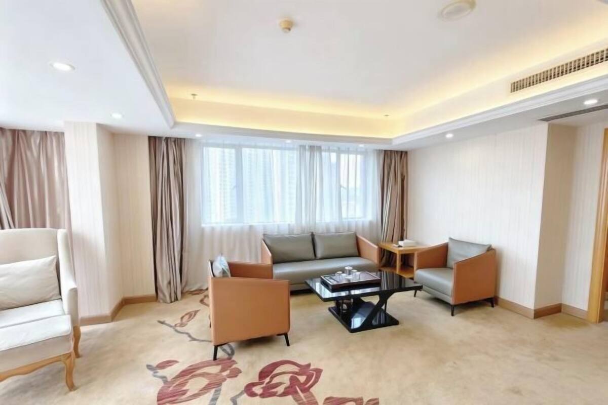 A modern hotel room with a sofa, armchairs, a coffee table, and a large window with curtains.