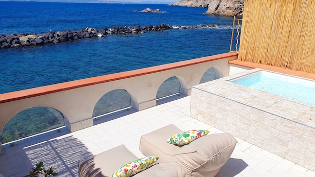 En tagterrasse med pool, to opholdsområder med blomsterpuder, samt udsigt til havet og kystlinjen.