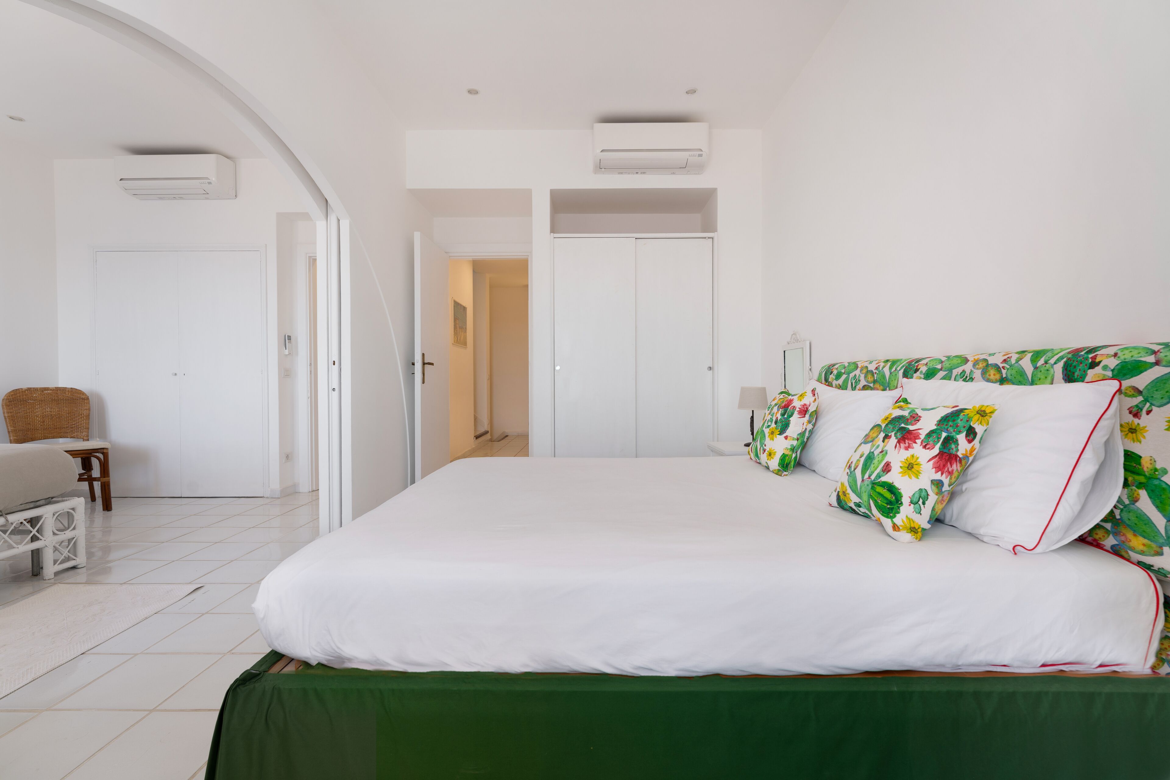 A bedroom with a bed, a green bedspread, white pillows, a wicker chair, and a white wardrobe.