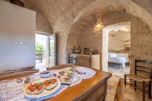 Cozinha rústica de pedra com mesa de madeira posta para refeição, cama e vista para o exterior.
