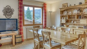 Un rincón comedor acogedor con una mesa y sillas de madera, un televisor y una estantería con vajilla y utensilios de cocina.