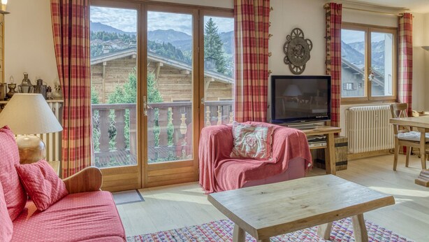 Un salón acogedor con un sofá, una mesa y un televisor. Amplias ventanas ofrecen vistas a montañas y una cabaña de madera.