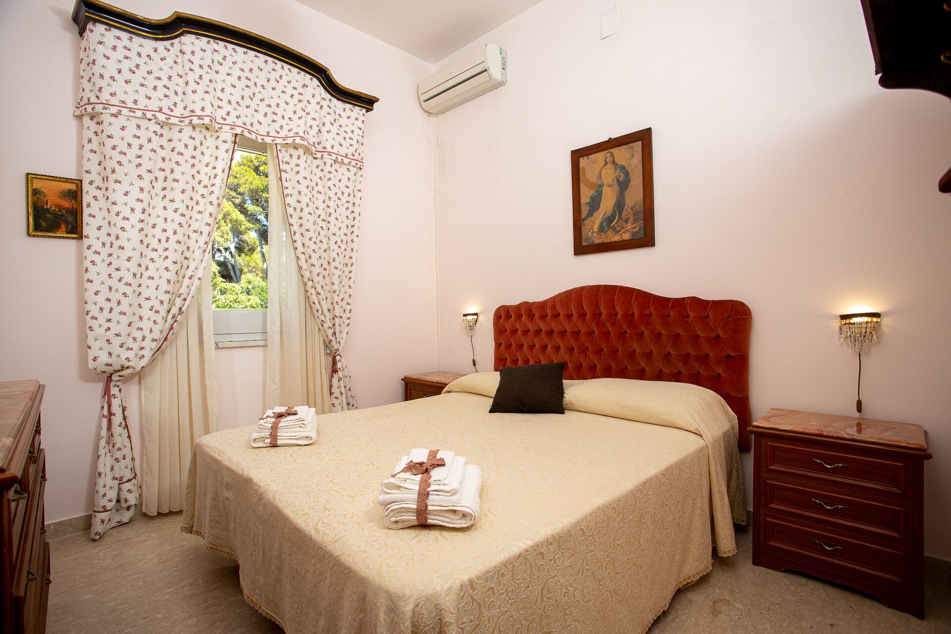 A bedroom with a large bed, a headboard, two bedside tables, a nightstand, a wooden dresser, a painting on the wall, and a window with curtains.