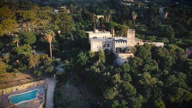 Vista aérea de uma estrutura semelhante a um castelo, cercada por vegetação densa, com uma piscina visível à esquerda.