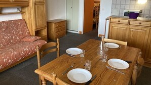 Une pièce chaleureuse avec une table à manger en bois, préparée pour deux personnes, un canapé et un coin cuisine doté de meubles en bois et d’un dosseret carrelé.