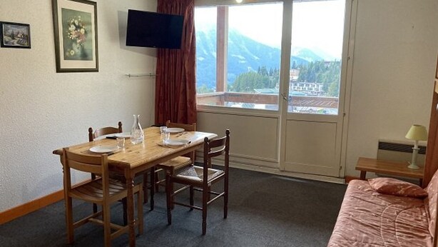 Une pièce comprenant une table à manger en bois, des chaises, un téléviseur, une fenêtre donnant sur les montagnes et un lit recouvert d’une couverture rose.