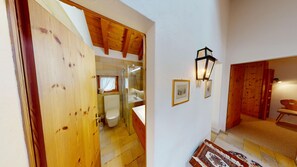 Una escalera de madera que lleva a un baño con cabina de ducha de vidrio, un lavabo blanco y cuadros enmarcados en la pared.