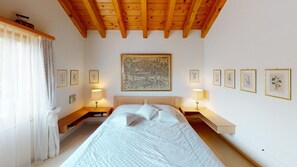 A bedroom with a wooden ceiling, a large bed with white bedding, two bedside tables with lamps, and framed artwork on the walls.
