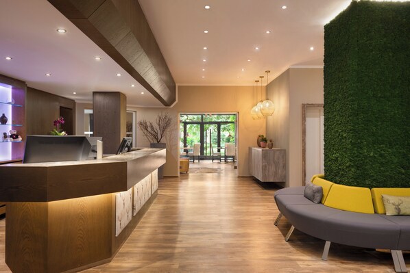 A reception area with a wooden reception desk, a seating area with a sofa, and a green wall feature. - City Hotel Siegen (Siegen)