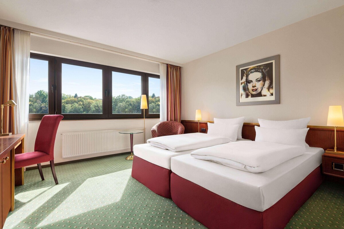 A hotel room with two beds, a desk, a chair, and a view of the outdoors.