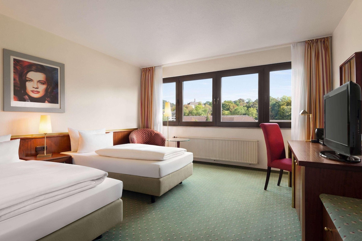 A hotel room with two beds, a desk with a television, a chair, a window with a view, and a painting on the wall.