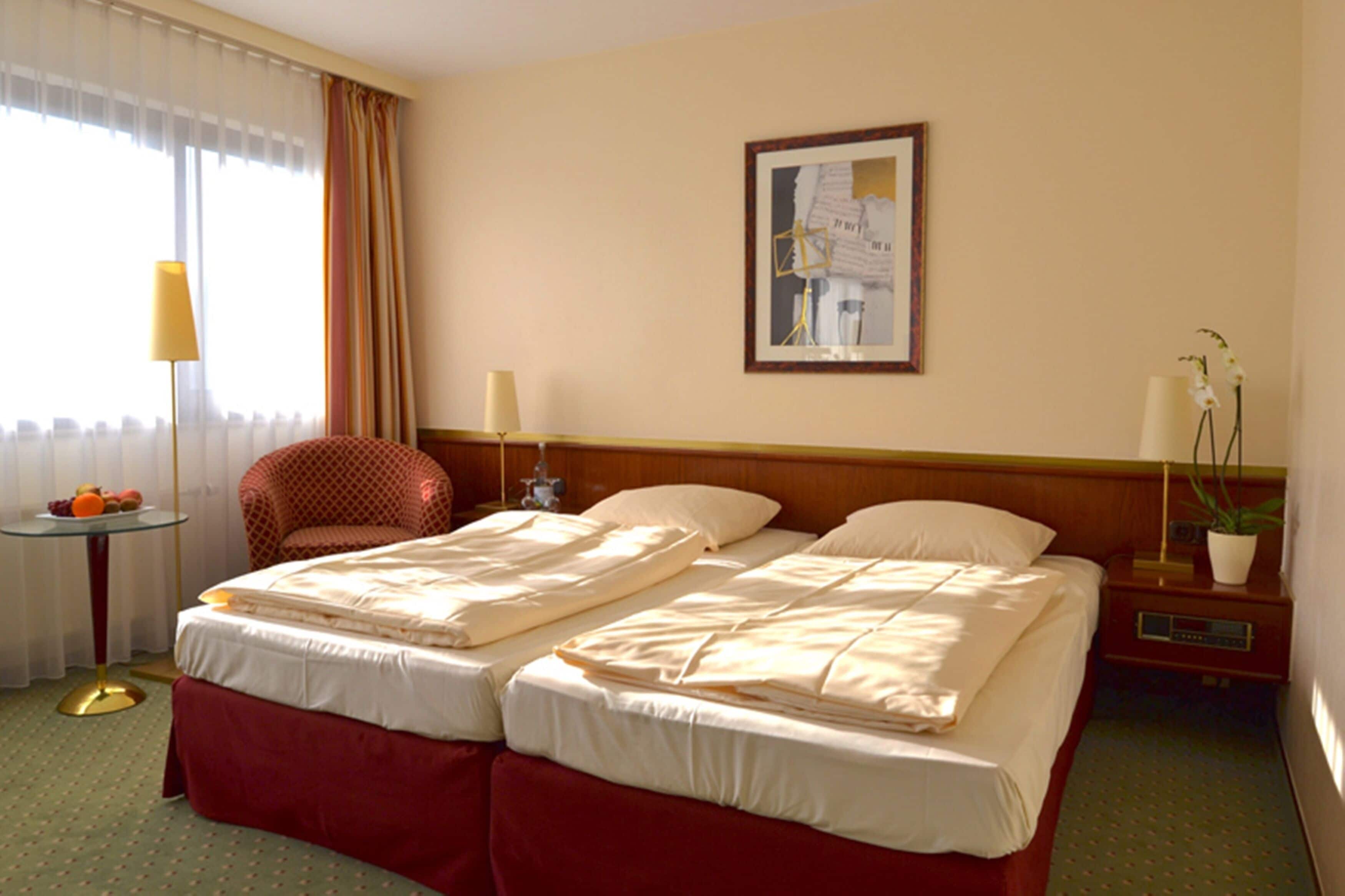 A hotel room with two beds, a chair, a table with fruit, a lamp, a framed picture on the wall, and a potted plant.