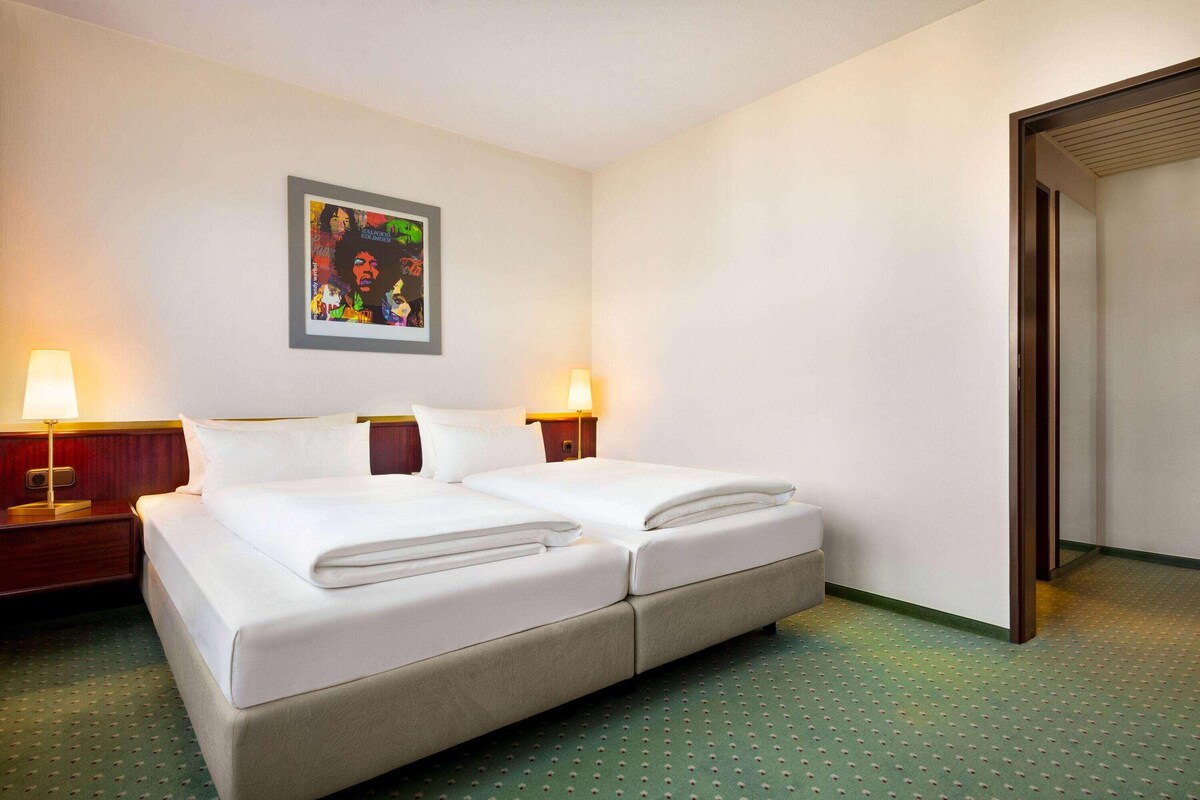 A hotel room with a double bed, two bedside tables with lamps, a framed artwork on the wall, and a green carpeted floor.