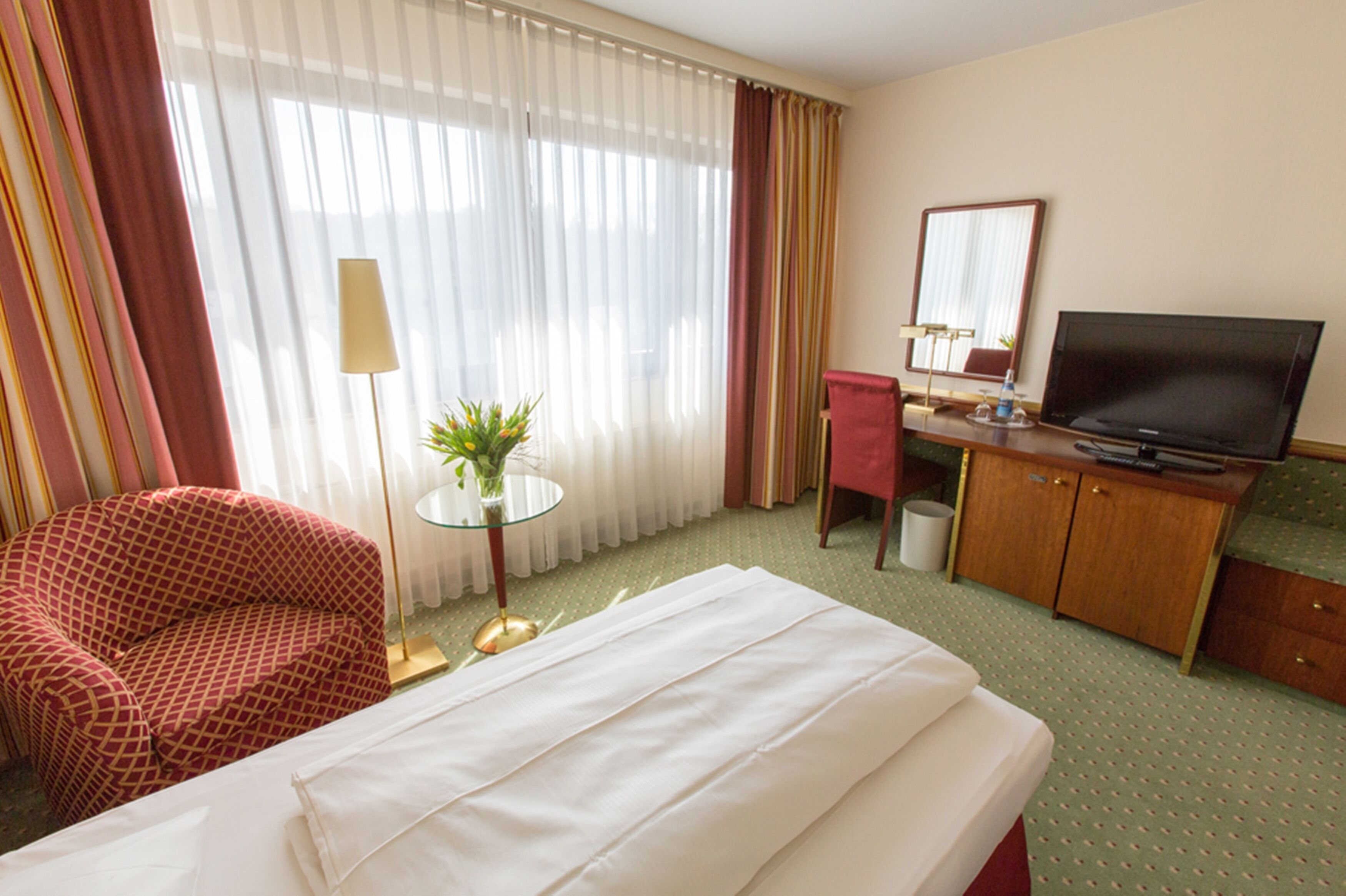 Ein Hotelzimmer mit einem Bett, einem Schreibtisch mit Fernseher, einem Sessel, einem kleinen Tisch mit Pflanze und einem Fenster mit Vorhängen.