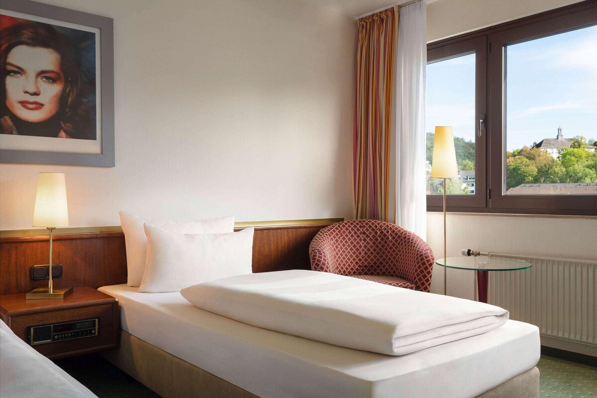 A hotel room with a bed, a nightstand with a lamp, a chair, a small table, and a window with a view of trees and buildings.